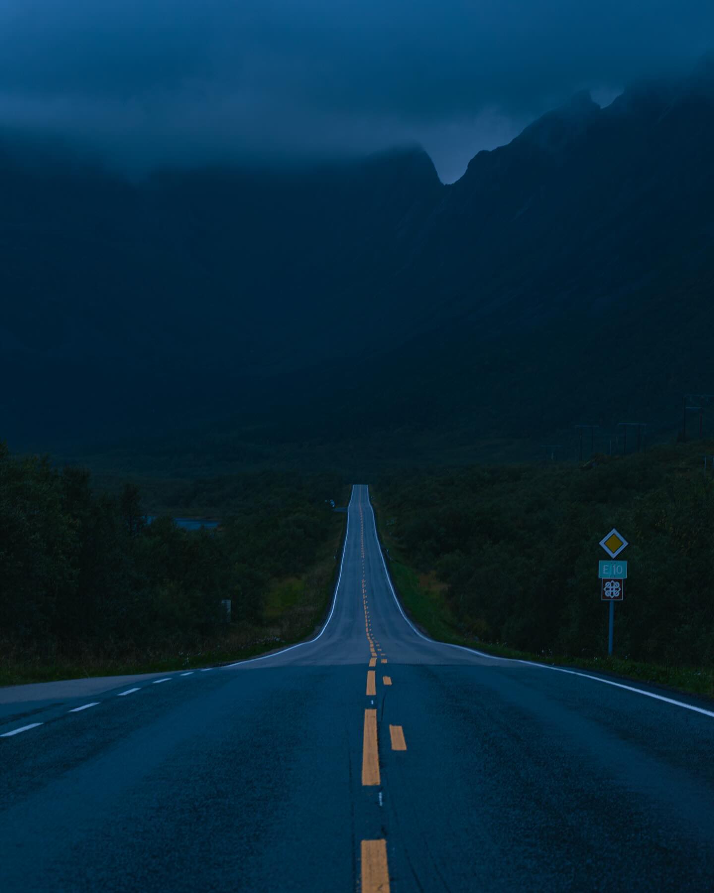 A long, empty road stretches into the distance under a dark, cloudy sky with mountains in the background.