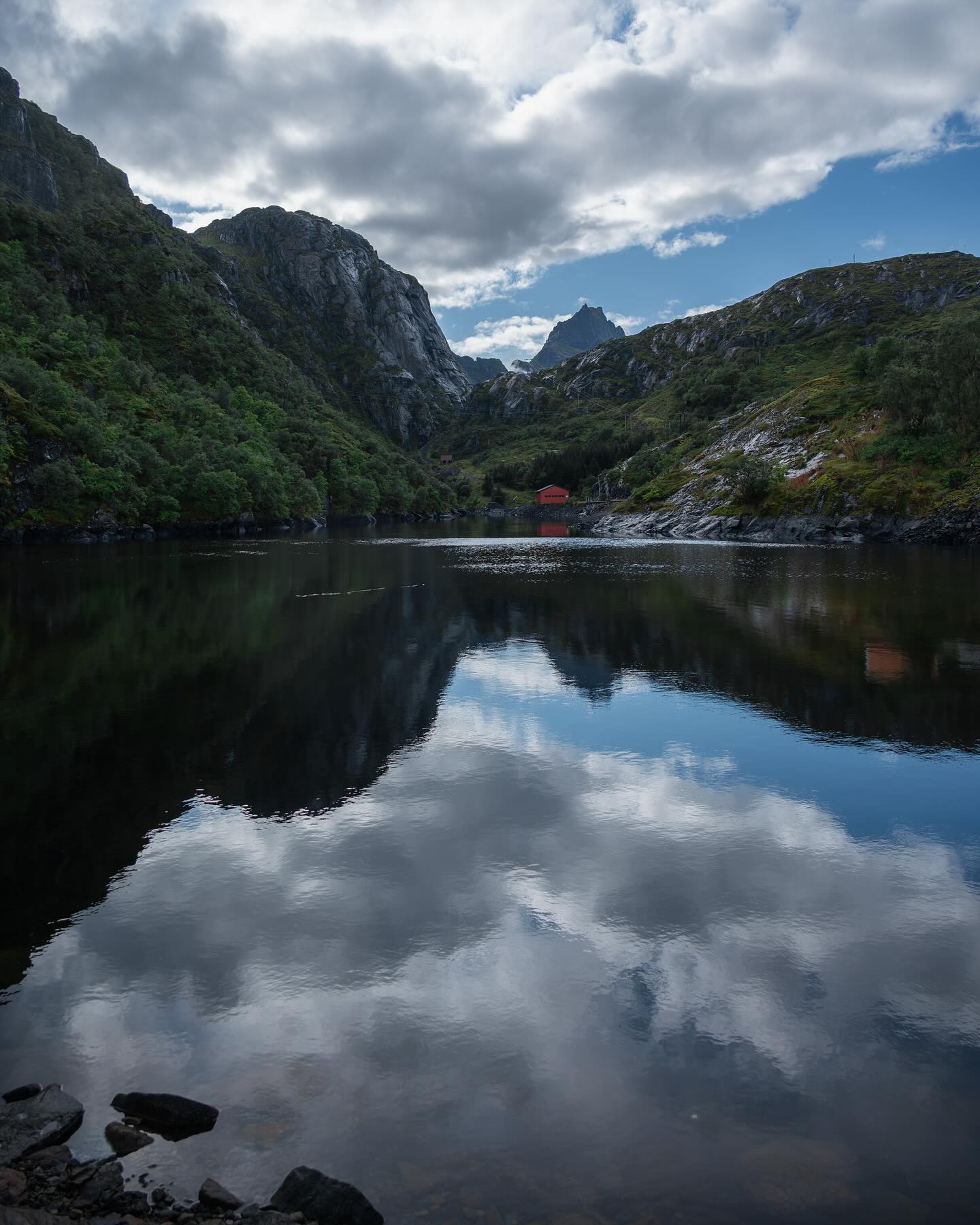 A calm lake reflects surrounding mountains and a partly cloudy sky. A small red cabin is nestled among green trees on the far shore.