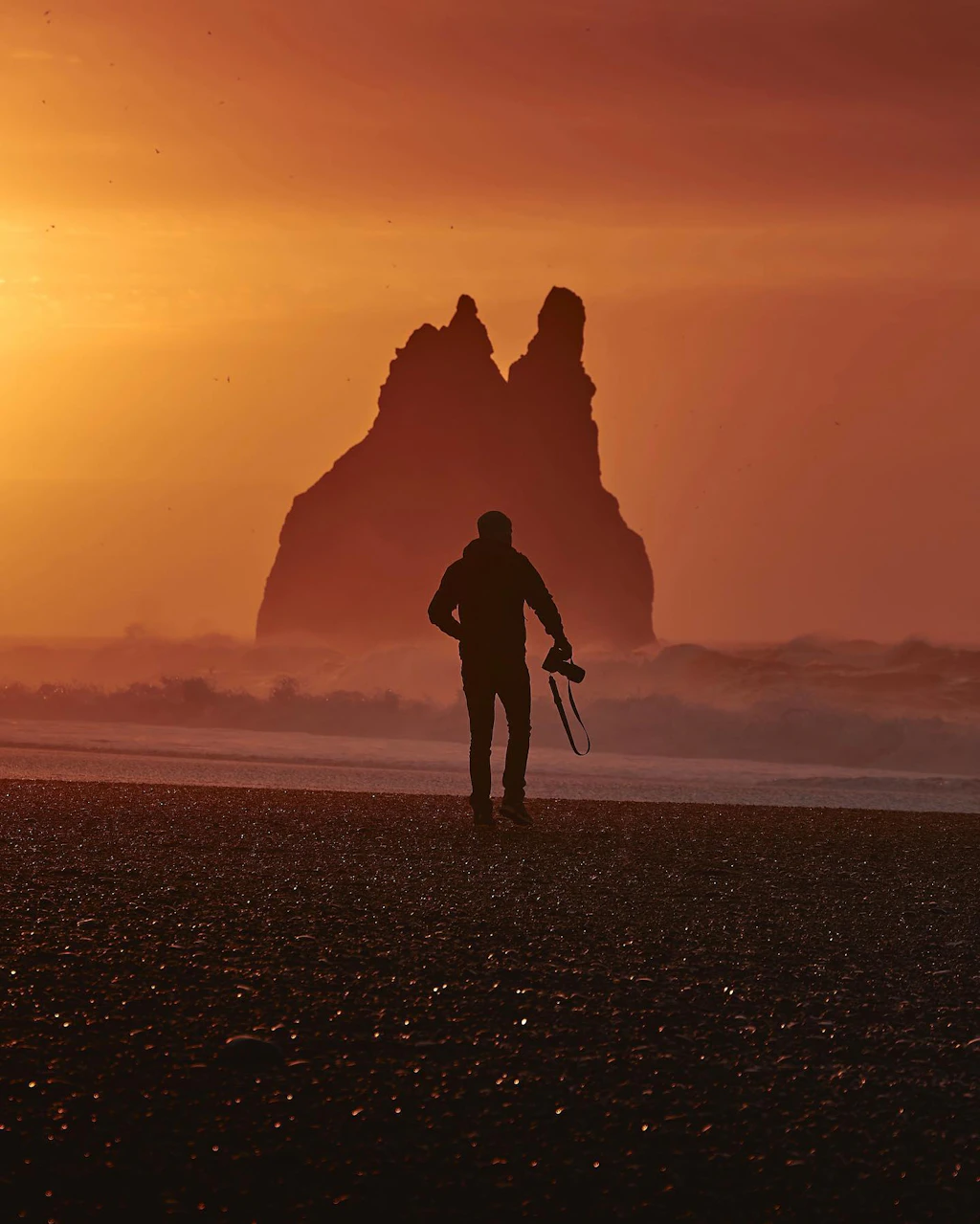 Silhouette of a man with a camera. He is on a beach with a sea stack in the distance. The sky is orange.